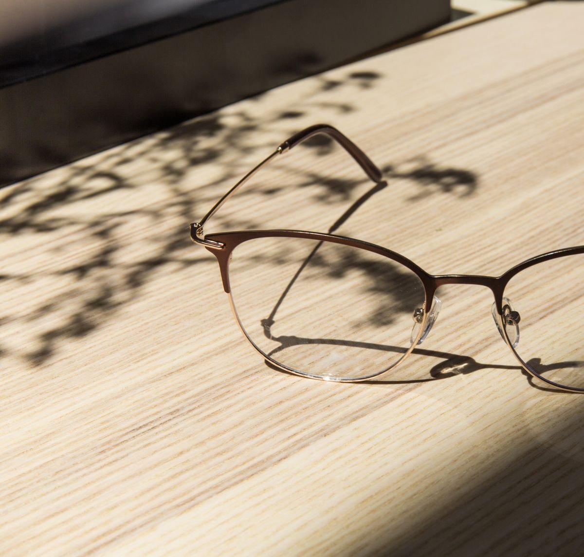 Unas gafas apoyadas sobre una mesa de madera clara, atravesada por la sombra blanda de unas hojas